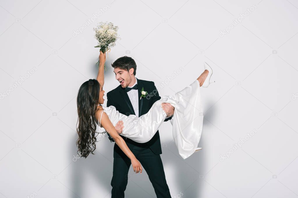 excited bridegroom holding happy african american bride on hands on white background