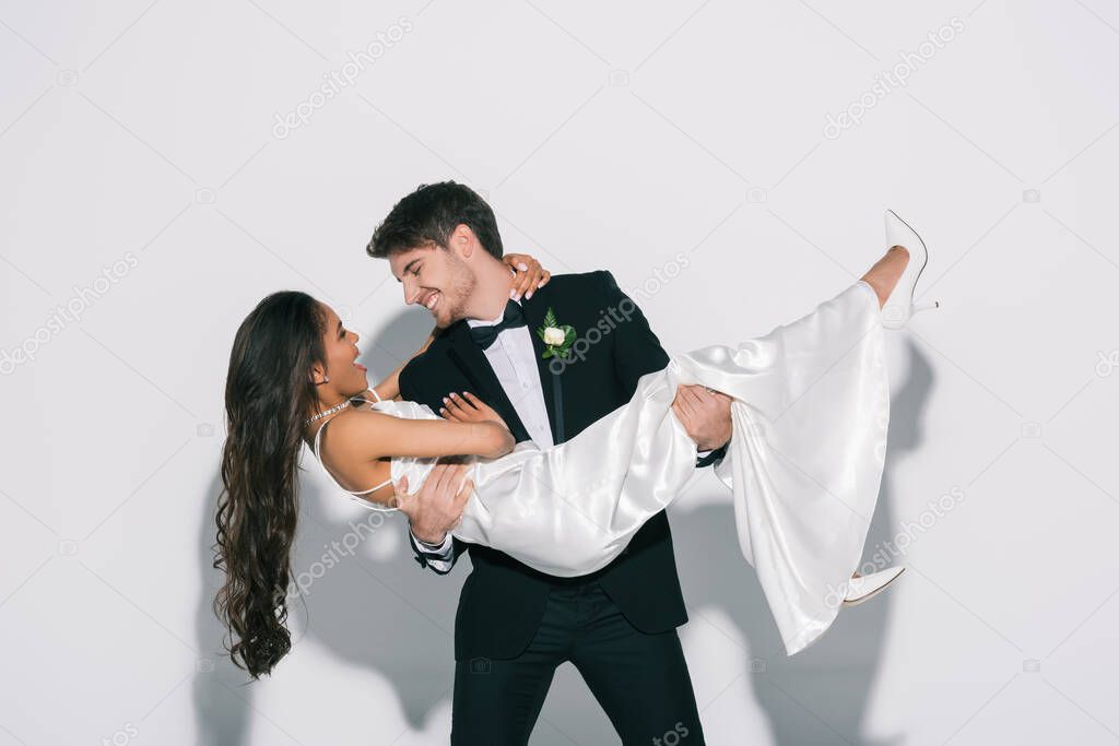cheerful bridegroom holding happy african american bride on hands on white background