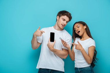 Yakışıklı erkek arkadaşı olan çekici Afrikalı Amerikalı kız mavi arka plandaki akıllı telefonları işaret ediyor.