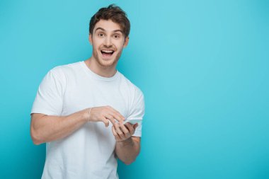 excited guy looking at camera while chatting on smartphone on blue background clipart