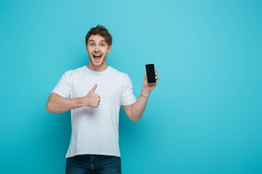 Heyecanlı genç adam mavi arka planda boş ekranlı akıllı telefonu tutarken baş parmağını gösteriyor.