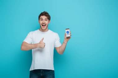 KYIV, UKRAINE - APRIL 23, 2019: excited young man showing thumb up while holding smartphone with Zoom app on screen on blue background clipart