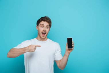 Heyecanlı adam mavi arka planda boş ekranlı akıllı telefonu işaret ediyor.
