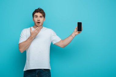 Mavi arka planda boş ekranlı akıllı telefonu gösterirken yüzünün yanında duran şok adam.