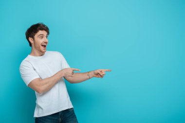 cheerful young man pointing with fingers while looking away on blue background clipart