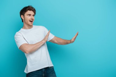 displeased young man showing refusal gesture while looking away on blue background clipart