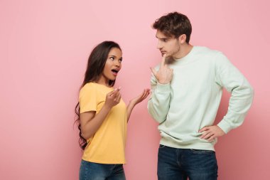 serious guy touching chin while looking at african american girlfriend quarreling on pink background clipart