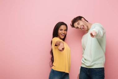 excited interracial couple pointing with fingers at camera on pink background