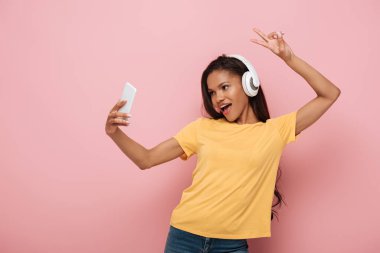 Kablosuz kulaklıklı mutlu Afro-Amerikalı kız akıllı telefondan selfie çekerken pembe arka planda zafer işareti gösteriyor.
