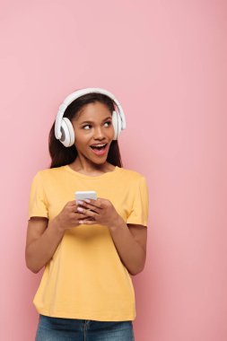 Pembe arka plana bakarken akıllı telefon kullanan kablosuz kulaklıklı neşeli Afrikalı Amerikalı kız.