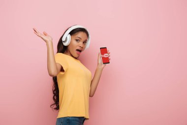 KYIV, UKRAINE - 23 Nisan 2019: pembe arka planda Youtube uygulaması ile akıllı telefonu gösteren kablosuz kulaklıklı heyecanlı Afrikalı Amerikalı kız