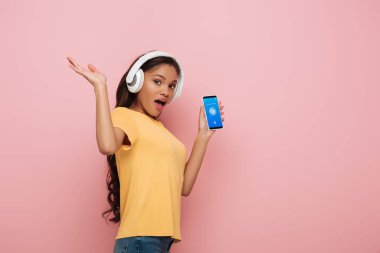 KYIV, UKRAINE - 23 Nisan 2019: Pembe arka planda Shazam uygulaması olan akıllı telefonu gösteren kablosuz kulaklıklı heyecanlı Afrikalı Amerikalı kız