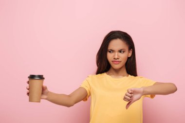displeased african american girl showing thumb down while holding coffee to go on pink background clipart