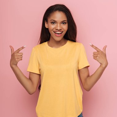 cheerful african american girl pointing with fingers at braces on her teeth on pink background clipart