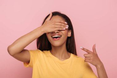 Neşeli Afro-Amerikalı kız elleriyle gözlerini kapatırken pembe dişlerindeki diş tellerini işaret ediyor.