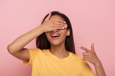 cheerful african american girl covering eyes with hand while pointing with finger at braces on her teeth isolated on pink clipart