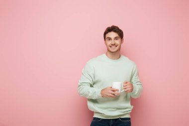 Pembe arka planda kameraya gülümserken beyaz bardak tutan yakışıklı genç adam.