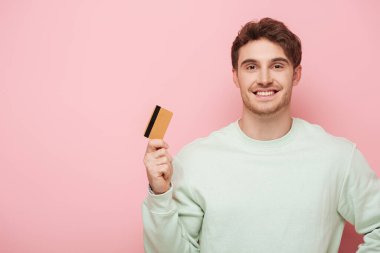 Pembe arka planda kameraya gülümserken elinde kredi kartı tutan mutlu genç adam.