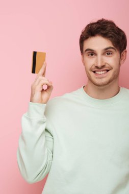 Pembe arka planda kameraya gülümserken elinde kredi kartı tutan neşeli genç adam.