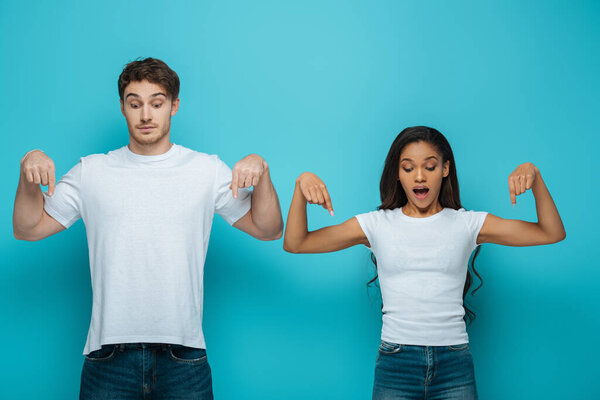 shocked interracial couple pointing with fingers and looking down on blue background 