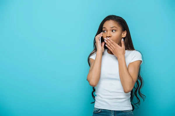 アフリカ系アメリカ人の女の子が手で口を覆い青で隔離されたスマートフォンで話しながら — ストック写真