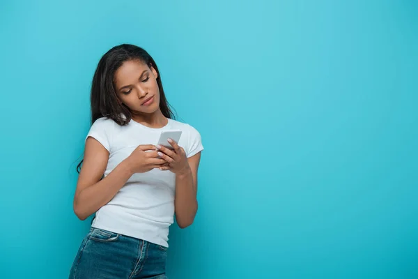 白いTシャツの魅力的なアフリカ系アメリカ人の女の子が青で隔離されたスマートフォンでチャット — ストック写真