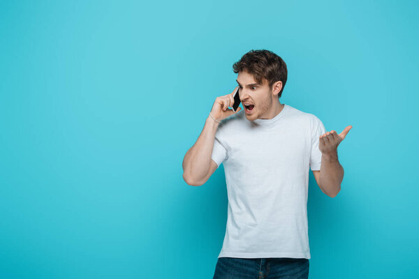 angry young man screaming on smartphone on blue background