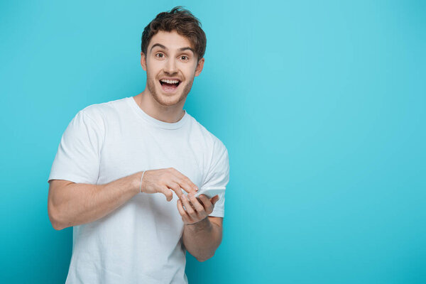 excited guy looking at camera while chatting on smartphone on blue background