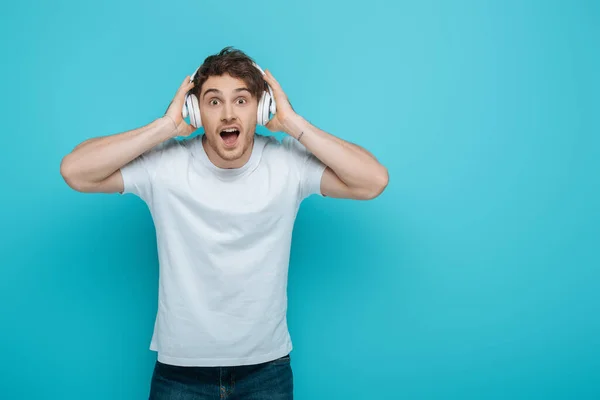 Shocked Young Man Touching Wireless Headphones While Looking Camera Blue — Stock Photo, Image