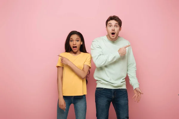 Shocked Interracial Couple Looking Camera Pointing Away Fingers Pink Background — Stock Photo, Image