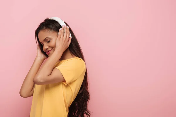 アフリカ系アメリカ人の女の子がピンクで隔絶された目でワイヤレスヘッドフォンで音楽を聴いて笑っています — ストック写真