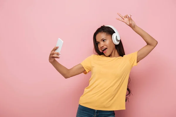 幸せなアフリカ系アメリカ人の少女がピンクの背景に勝利サインを見せながらスマートフォンで自撮り — ストック写真