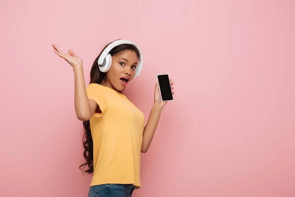 兴奋的非洲女孩在无线耳机显示智能手机与粉红背景的空白屏幕 — 图库照片