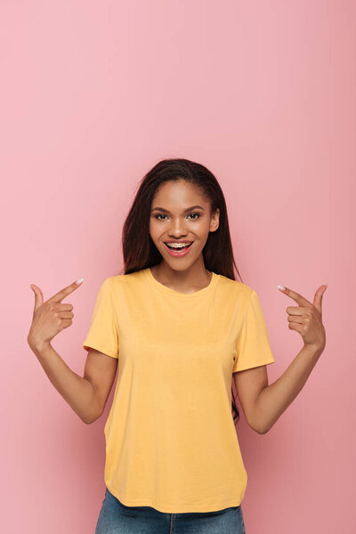 улыбающаяся африканская американка, указывающая пальцами на брекеты на розовом фоне
