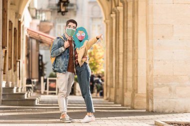 Tıbbi maskeli iki turist sokakta sarılıyor ve parmaklarıyla işaret ediyor.