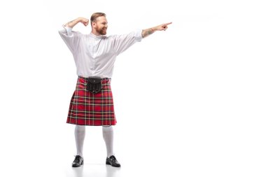happy Scottish redhead man in red kilt pointing with finger aside on white background clipart