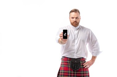 KYIV, UKRAINE - 29, 2019: kırmızı kiltli ciddi İskoç kızıl saçlı adam beyaz üzerine izole edilmiş tiktak uygulamalı akıllı telefon sunuyor.