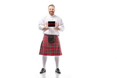 KYIV, UKRAINE - NOVEMBER 29, 2019: kırmızı kiltli gülümseyen İskoç kızıl adam beyaz arka planda Netflix uygulaması ile dijital tablet sunuyor