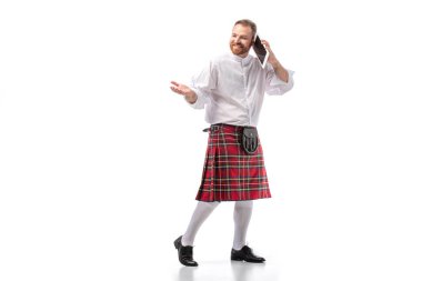 smiling Scottish redhead man in red kilt talking on digital tablet on white background clipart
