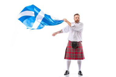 Scottish redhead man in red kilt with flag of Scotland on white background clipart
