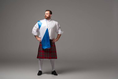 serious Scottish redhead man in red kilt with flag of Scotland and hands on hips on grey background clipart