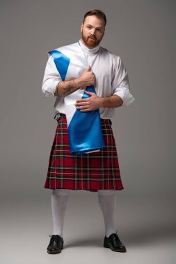 Scottish redhead man in red kilt with flag of Scotland on grey background clipart