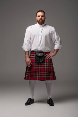 Scottish redhead man in red kilt with hand on hip on grey background clipart