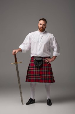 Scottish redhead man in red kilt with sword on grey background clipart