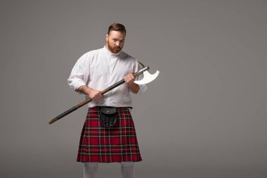serious Scottish redhead man in red kilt with battle axe on grey background clipart