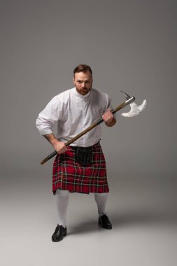 Gri arka planda savaş baltası olan kırmızı kiltli kızgın İskoç adam.