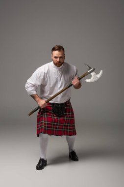 angry Scottish redhead man in red kilt with battle axe on grey background clipart