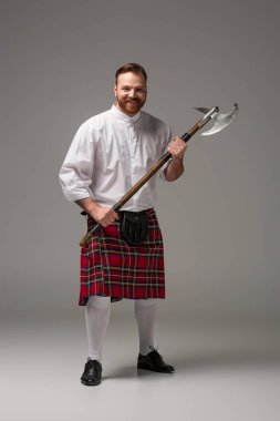 Gri arka planda savaş baltası olan kırmızı kiltli, gülümseyen İskoç kızıl adam.