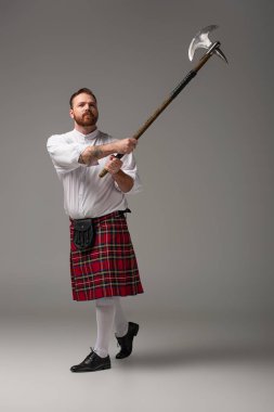 Scottish redhead man in red kilt raising battle axe on grey background clipart