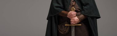 cropped view of medieval Scottish knight in mantel with sword in hands on grey background, panoramic shot clipart